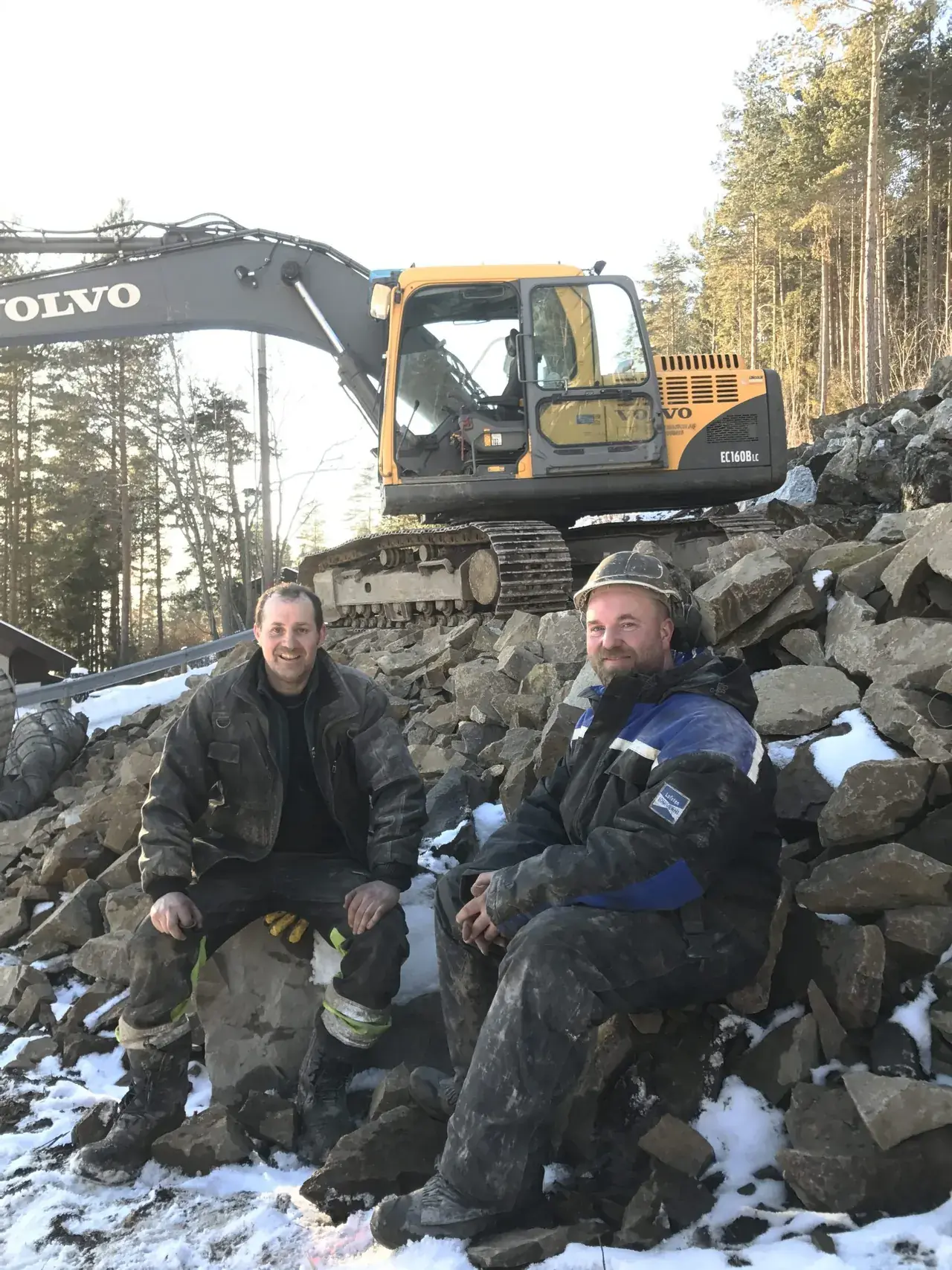 Rune Antonsen og Vebjørn Grimnes sitter på sprengstein foran gravemaskinen.