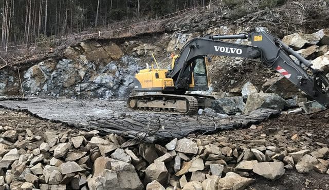 Gravemaskin på sprengningsmatter foran en fjellskjæring.