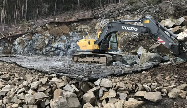 Gravemaskin på sprengningsmatter foran en fjellskjæring.