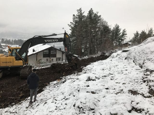 Gravemaskin graver ut en snødekt skråning ved et hus.