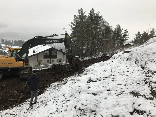 Gravemaskin graver ut en snødekt skråning ved et hus.